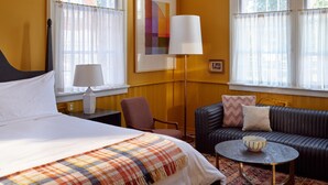 A hotel room with a bed, a sofa, a small table, a floor lamp, and two windows with curtains. - The Abner Hotel (Litchfield)