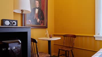 Quarto de hotel com uma cadeira de madeira, uma mesa redonda pequena, um quadro na parede e um rádio num armário.