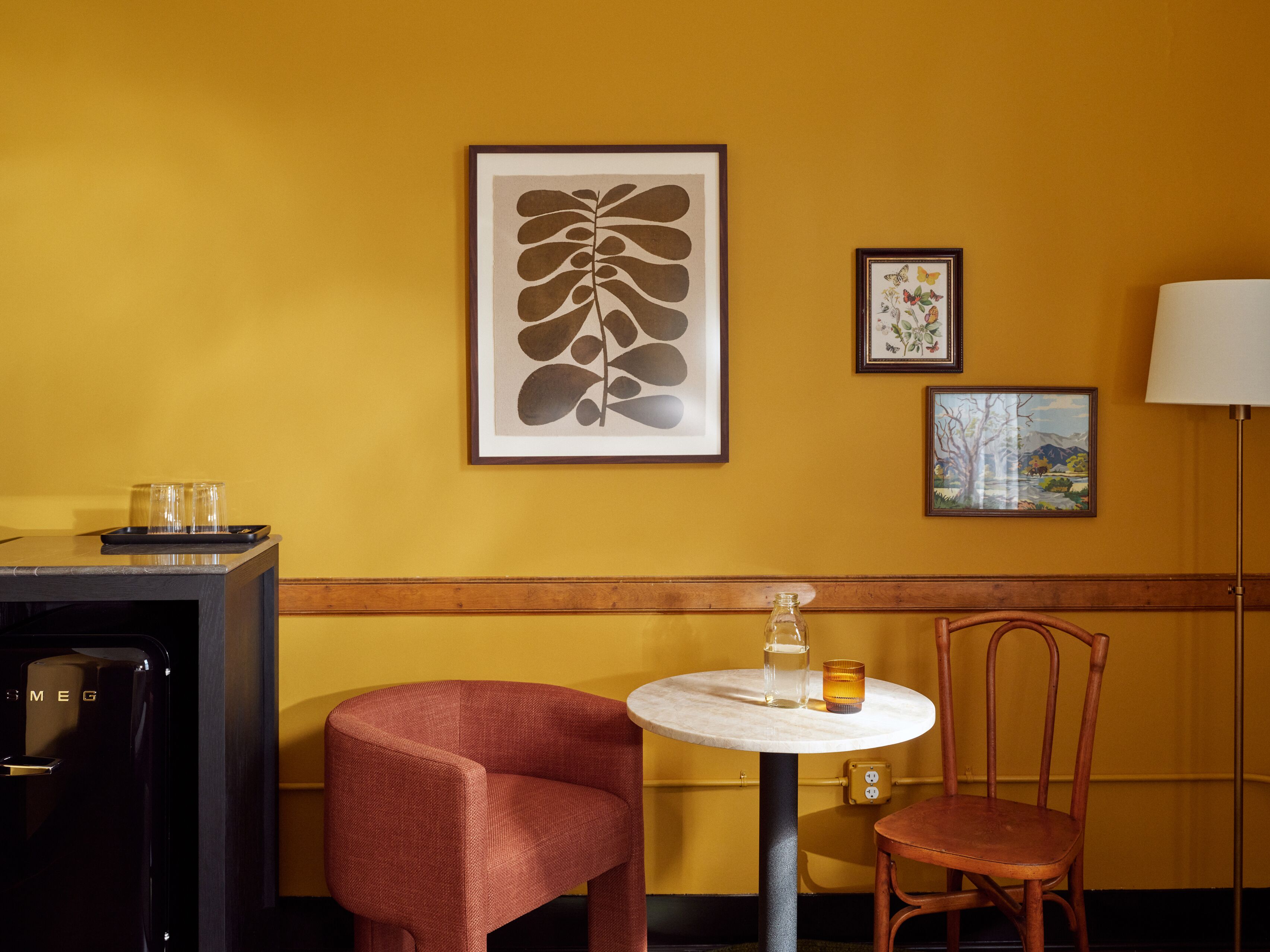 Una habitación con una pared amarilla, una obra de arte con una hoja enmarcada, una obra de arte con una mariposa enmarcada, una mesa redonda con una botella y un vaso, una silla roja y una lámpara.