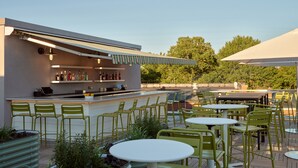 An outdoor seating area with green chairs and tables, a bar with shelves of bottles, and a view of trees in the background. - The Abner Hotel (Litchfield)