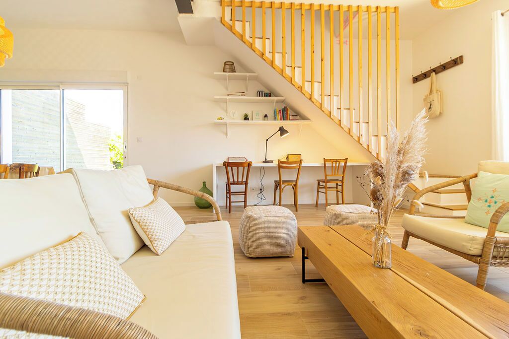 A modern living room with a staircase, a sofa, chairs, a wooden table, and a desk with a lamp.