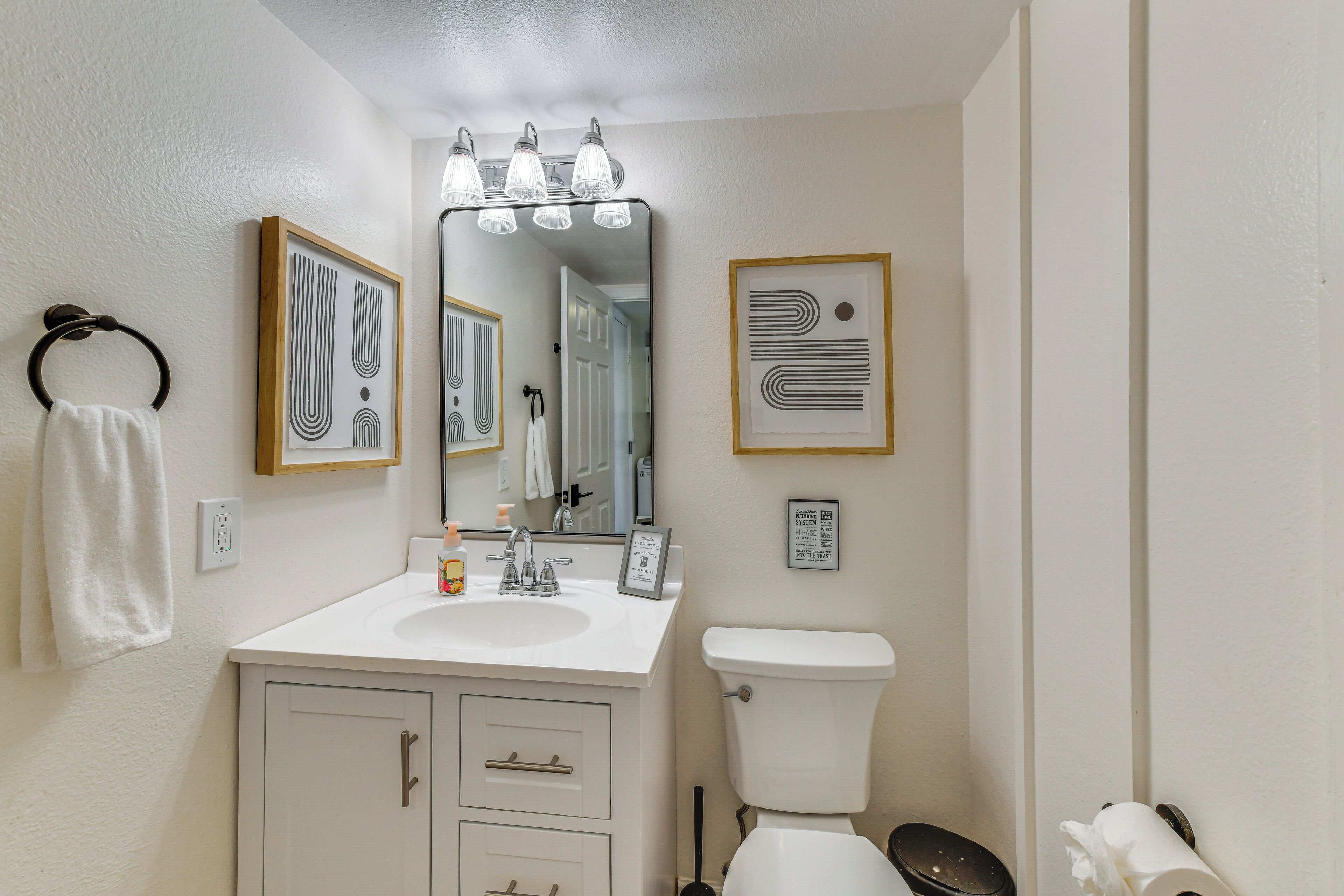 A bathroom with a white sink, a mirror, a toilet, and framed artwork on the wall.