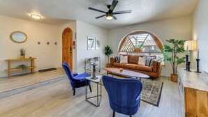 Un salon moderne avec un plancher en bois, un ventilateur de plafond, un canapé en cuir marron, des fauteuils bleus, une table basse et une grande fenêtre donnant sur un beau paysage.