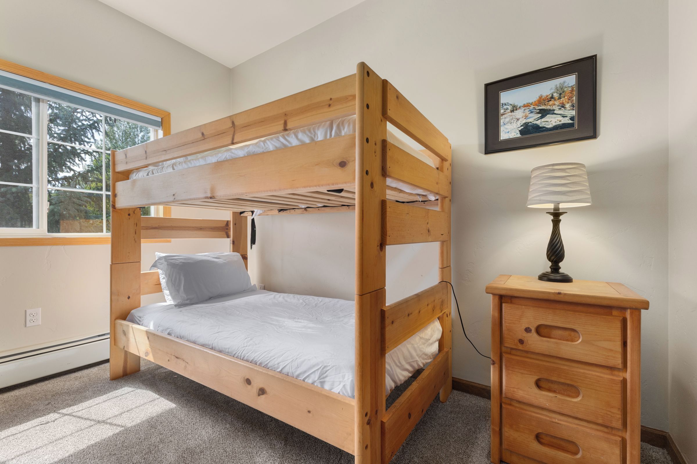 A bunk bed with a wooden frame, a bedside table with drawers, a lamp, and a framed picture on the wall.