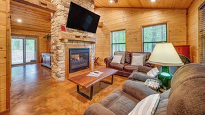 A cozy cabin interior with a stone fireplace, wooden walls, a leather sofa, and a flat-screen TV mounted above the fireplace. - Broken Bow Cabin: Hot Tub, Fire Pit & 7 Mi to Lake (Broken Bow)