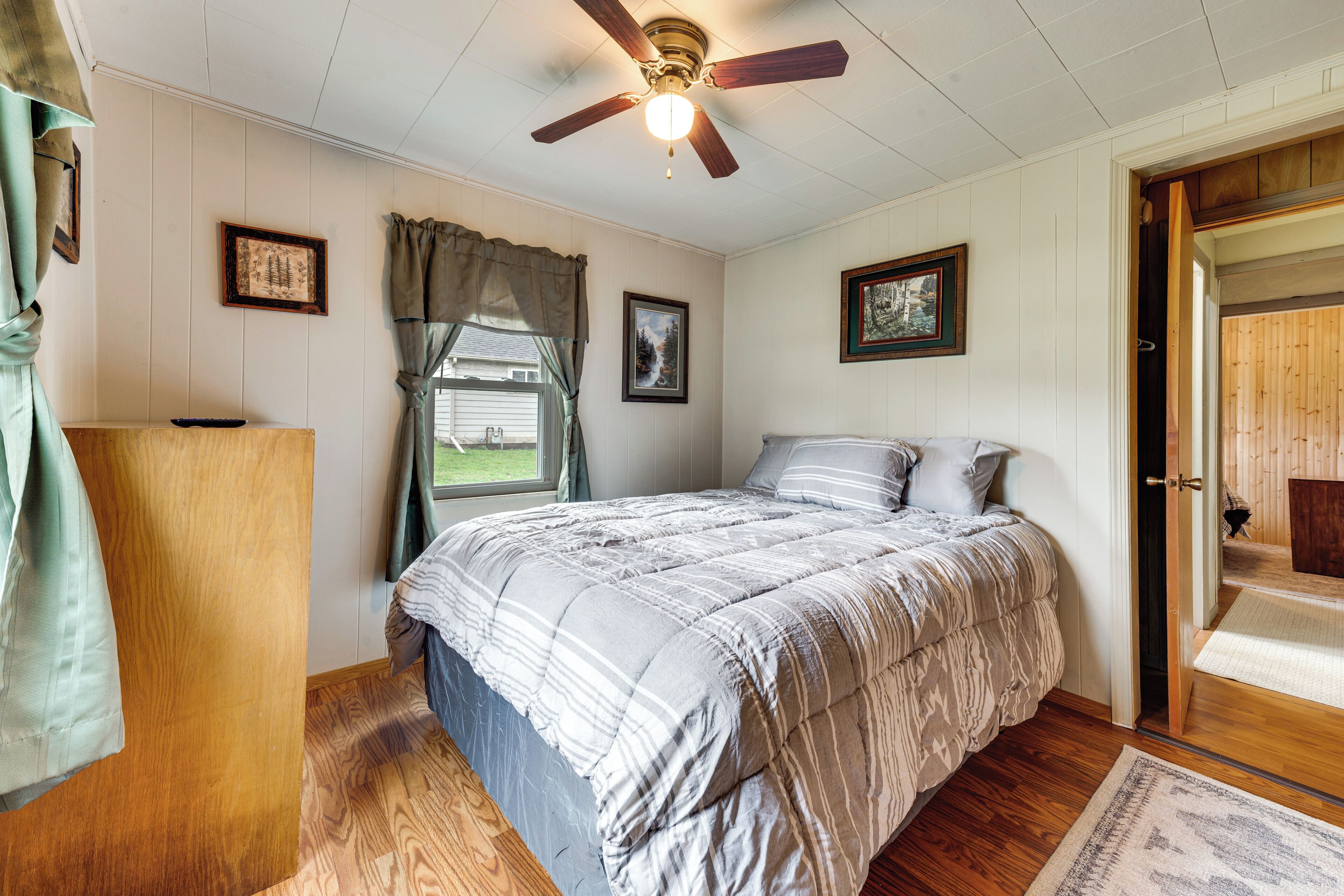Un dormitorio con cama, piso de madera, ventilador de techo y una puerta que da a otra habitación.