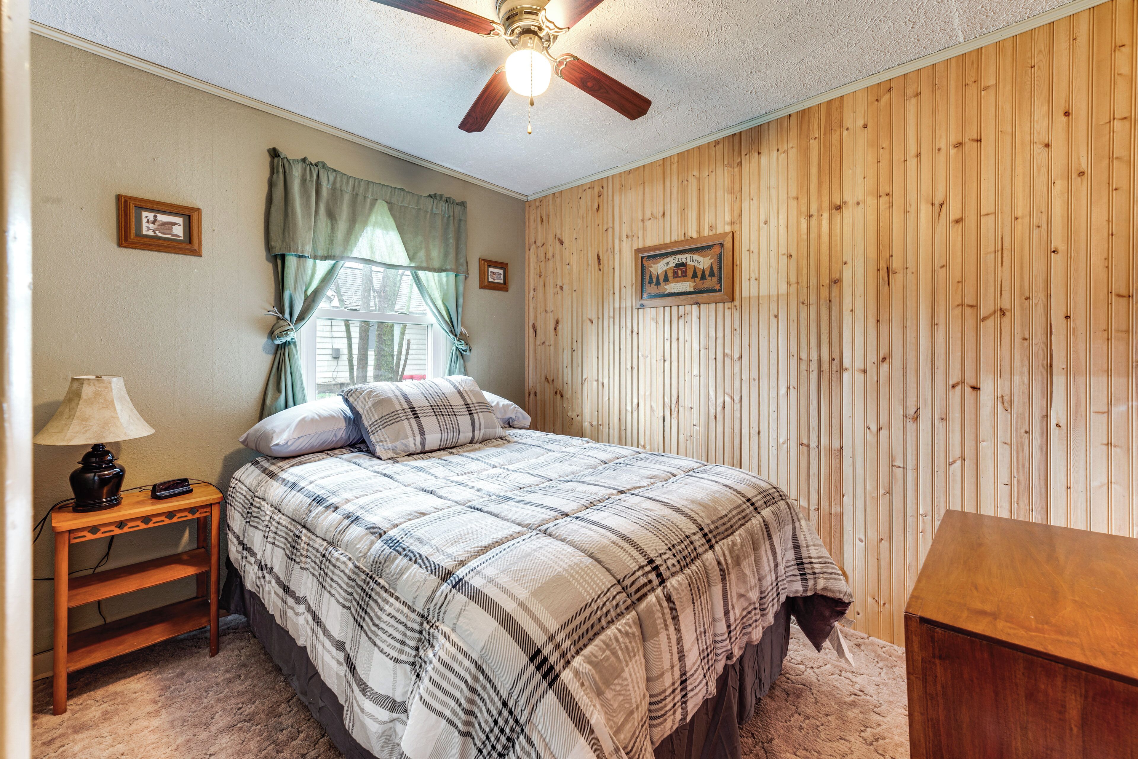 A bedroom with a bed, a nightstand, a lamp, a ceiling fan, and wooden paneling on the walls.