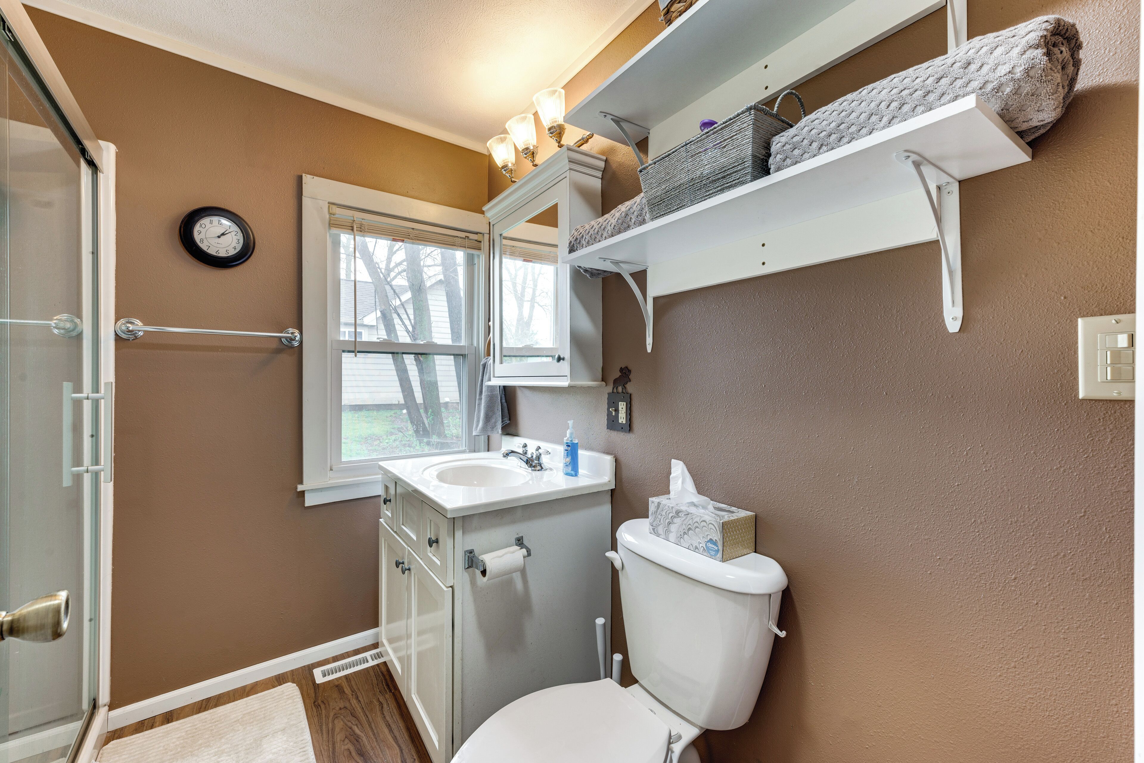 A bathroom with a toilet, sink, and shower. There are shelves above the toilet and a clock on the wall.