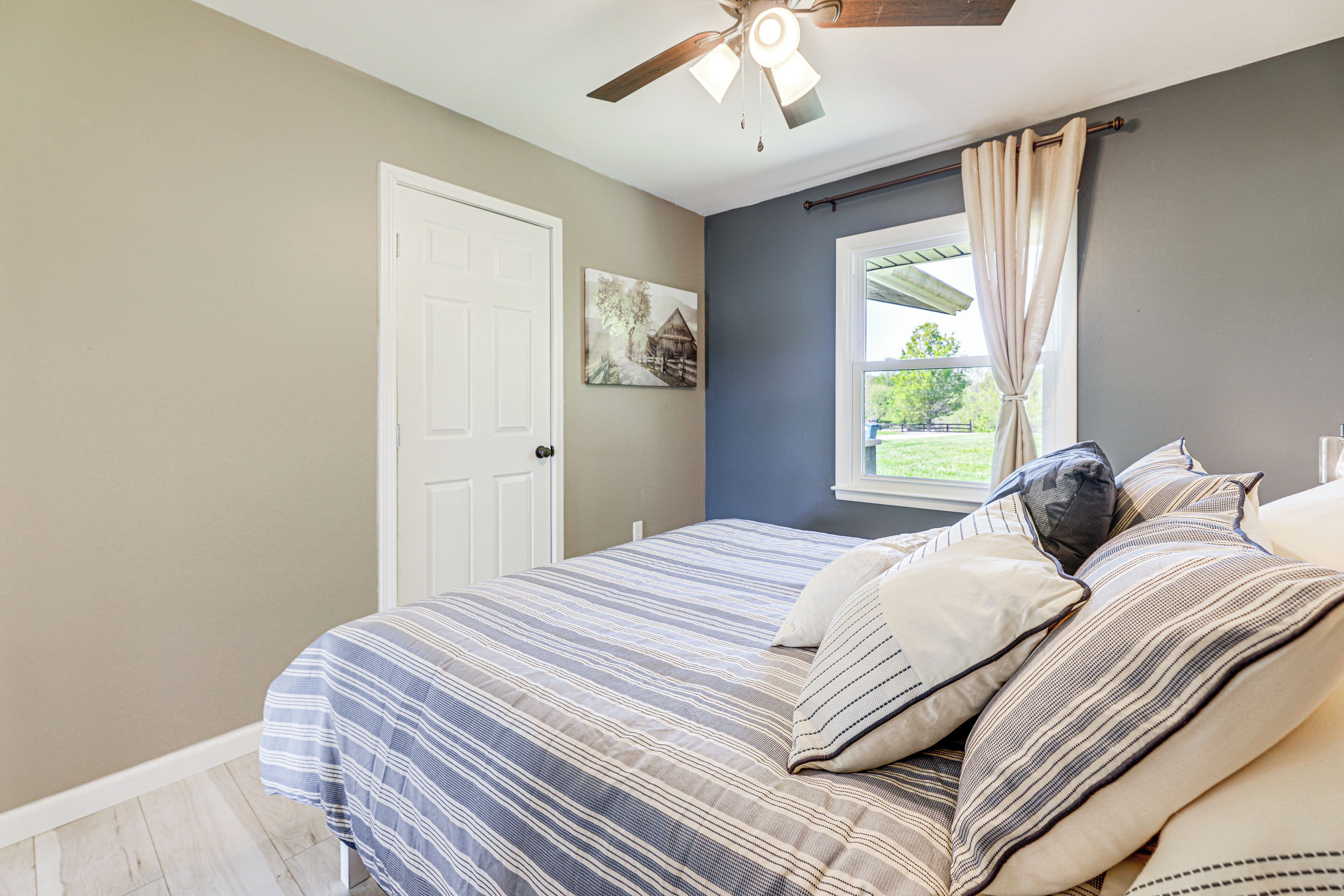 Um quarto com cama, ventilador de teto, janela com cortinas e porta.