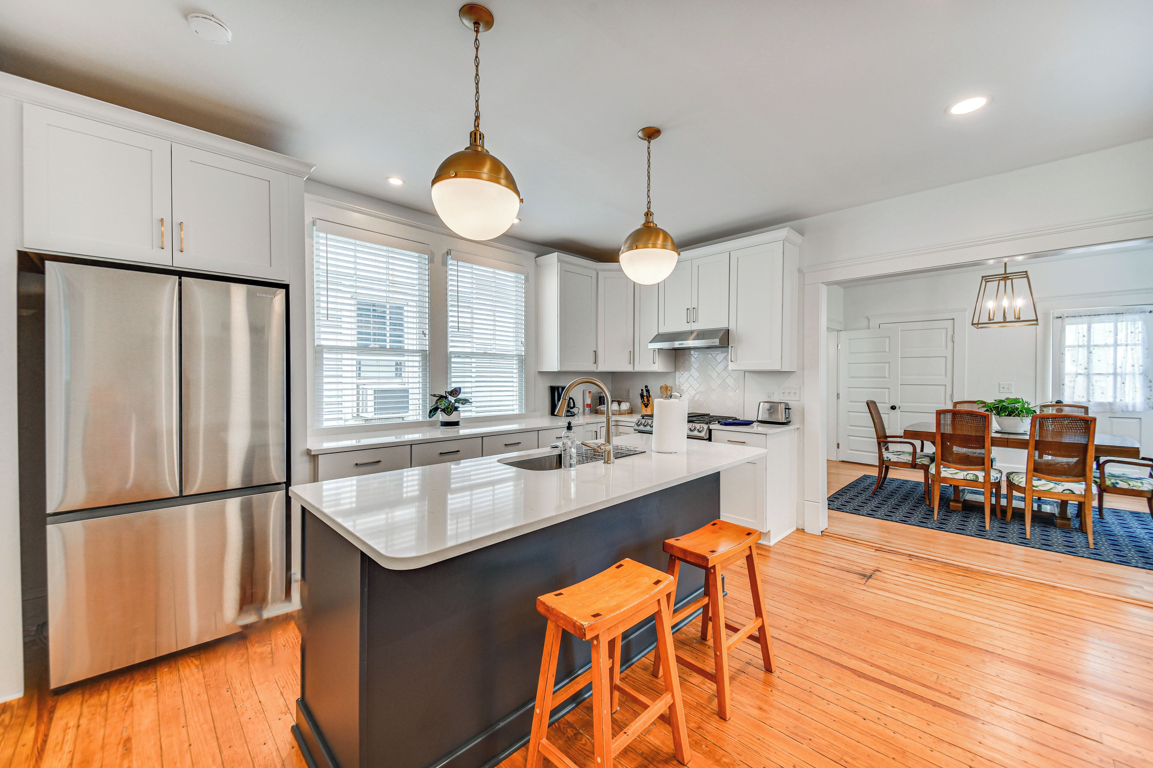 A modern kitchen with stainless steel appliances, a central island with stools, and a dining area with wooden chairs and a blue rug.