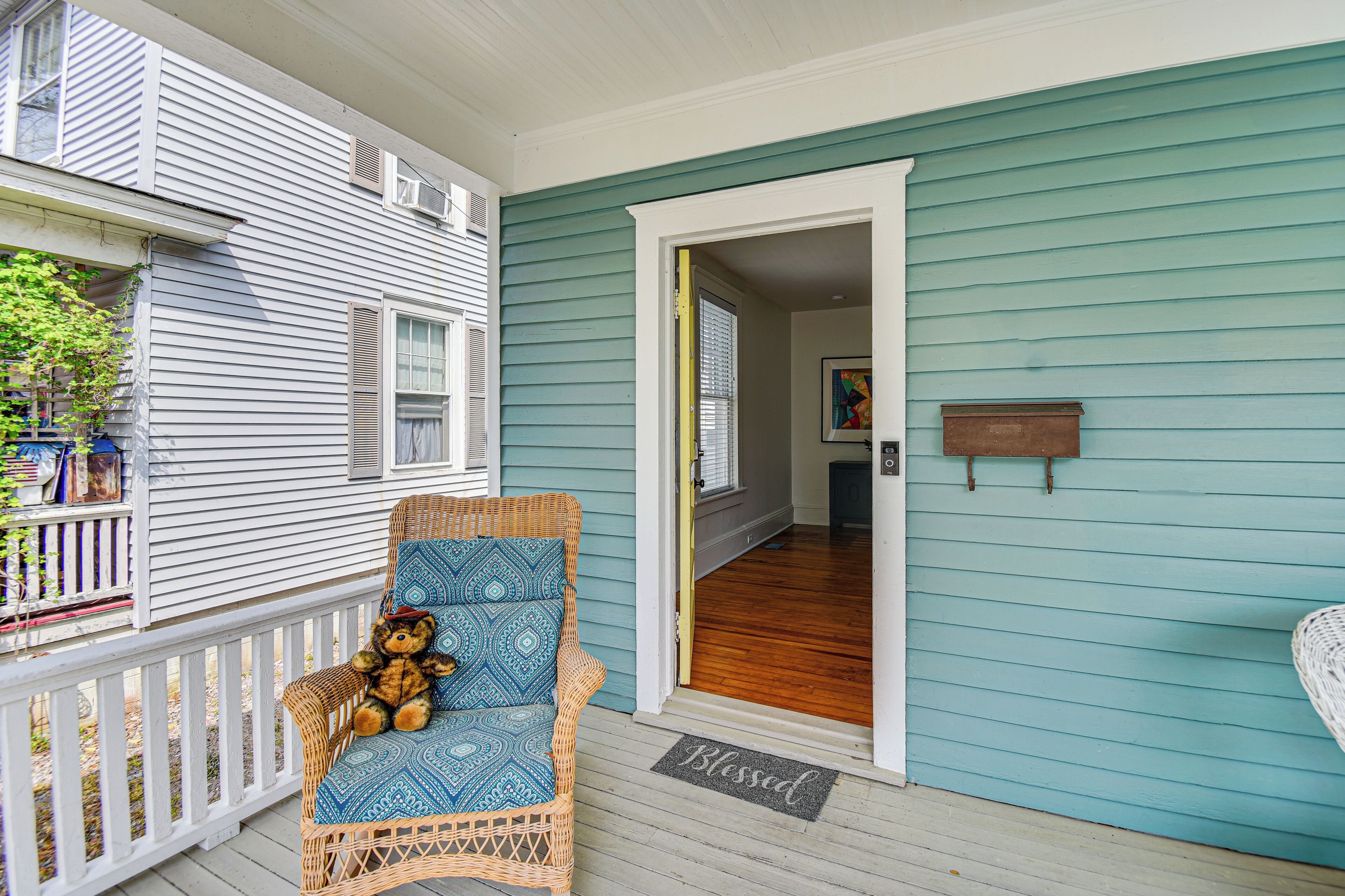 A porch with a wicker chair and a teddy bear, a wooden door, and a doormat.