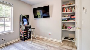 Una habitación con una televisión de pantalla plana en la pared, una consola Nintendo Switch, una estantería con libros y juegos, y un ventanal que proporciona luz natural.