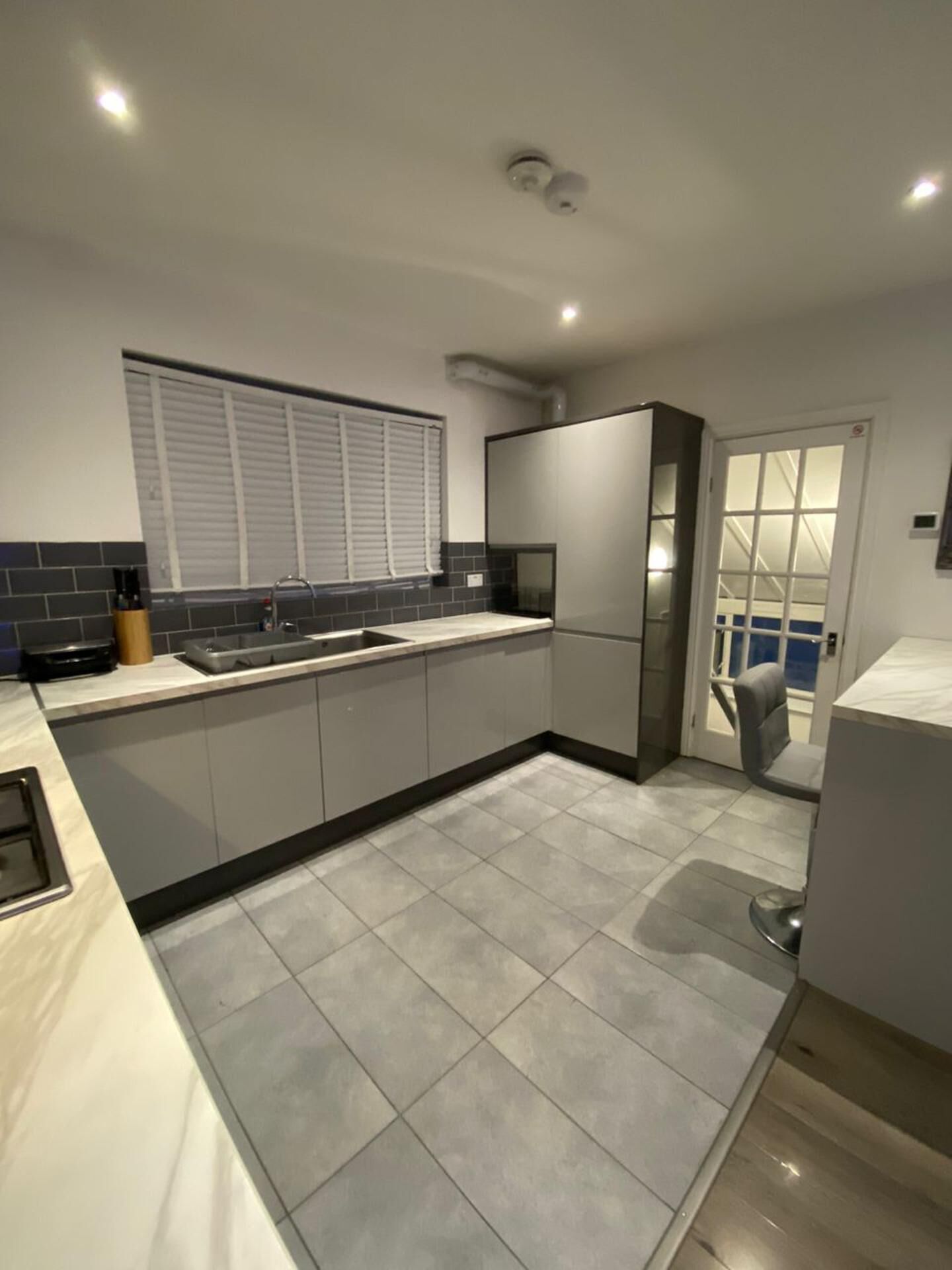 A modern kitchen with grey tiles, a built-in sink, and a window with blinds.