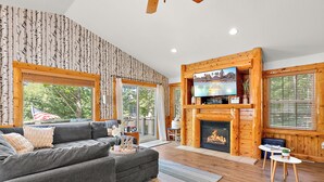 A cozy living room with a fireplace, a grey sofa, and a television. - The Treehouse @ Higgins Lake-HOT TUB- Fireplace-Pets❤️-3 min walk to the lake (Roscommon)
