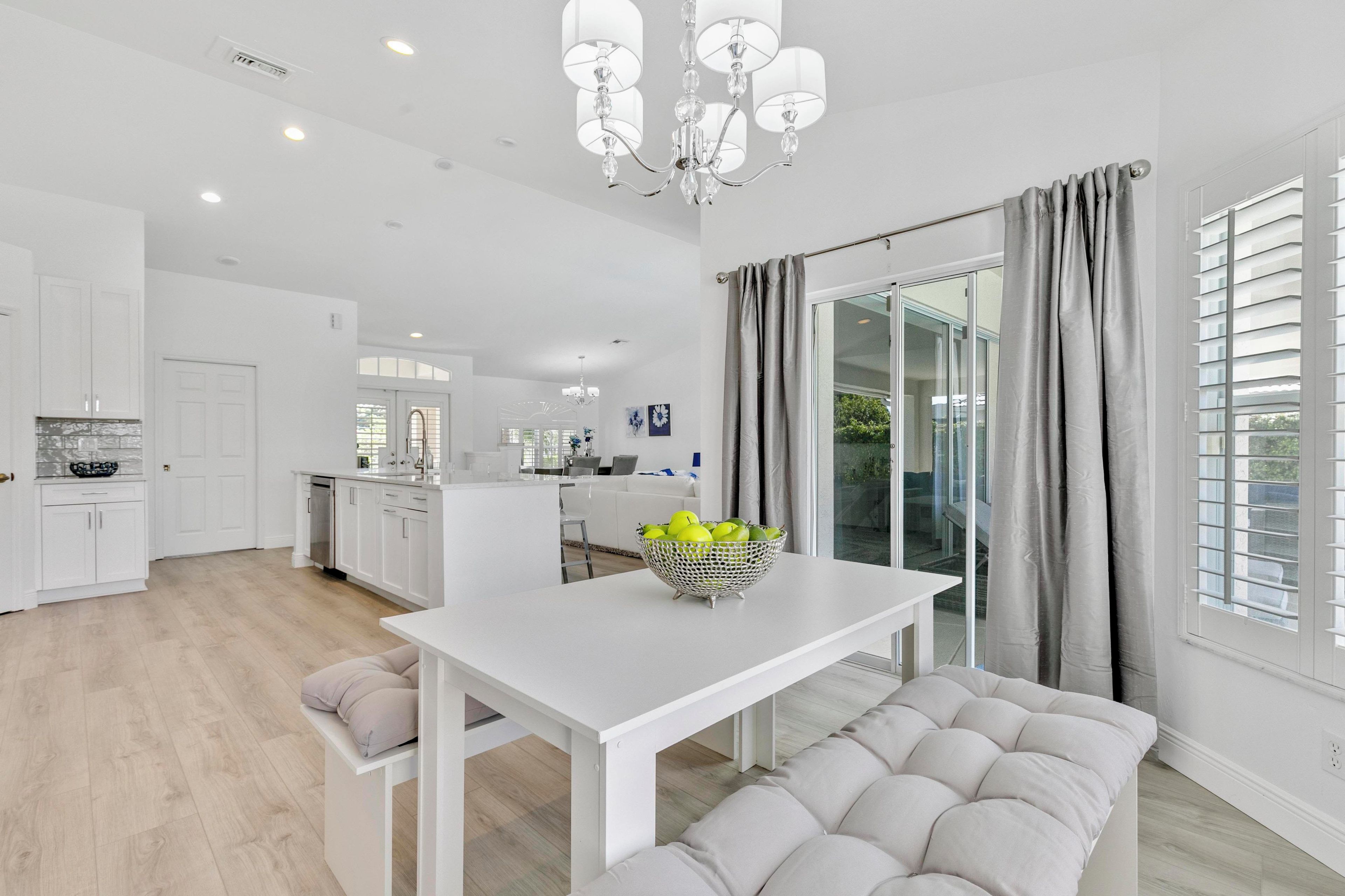 A modern kitchen with white cabinetry, a dining area with a white table and bench, and a large window with grey curtains.