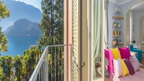 A balcony with a view of a lake and mountains, a bed with colorful pillows, and a room with a bookshelf and a mirror. - Villa Artemisia by MyVillaCollection (Lenno)
