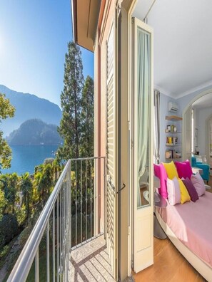 A balcony with a view of a lake and mountains, a bed with colorful pillows, and a room with a bookshelf and a mirror. - Villa Artemisia by MyVillaCollection (Lenno)