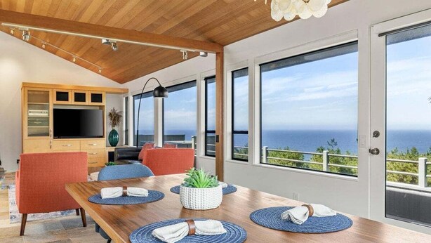 Un espace repas moderne avec une table en bois, des sets de table bleus et une vue sur l’océan grâce à de grandes fenêtres.