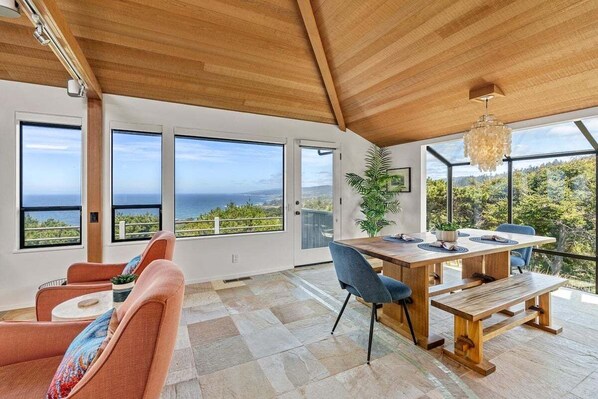 A spacious room with a wooden ceiling, large windows offering ocean views, a dining area with a wooden table and chairs, and a seating area with a sofa and armchairs.