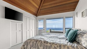 Une chambre avec un grand lit, un plafond en bois, un téléviseur fixé au mur et une vue sur l’océan par de grandes fenêtres.