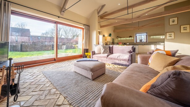 A spacious living room with a large sliding glass door leading to a backyard, a cozy seating area with a sofa and ottoman, and a kitchen in the background. - Harnser Barn, Tittleshall, Norfolk (Tittleshall)