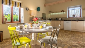 A kitchen with a dining area, wooden beams, a window with a rooster decoration, and a potted plant on the windowsill. - Harnser Barn, Tittleshall, Norfolk (Tittleshall)