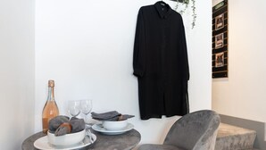 Een eethoek met een tafel gedekt voor twee personen, een fles wijn en een schaal fruit. Een zwart jasje hangt aan de muur, en er staat een grijze stoel.