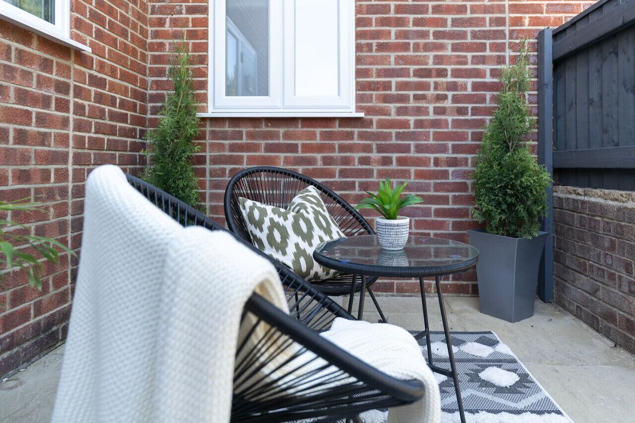 A small patio area with a black wicker chair, a round glass table, and two potted plants.