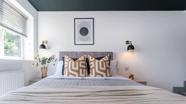 Una cama bien hecha con almohadones estampados, una obra enmarcada encima y lámparas fijadas a la pared a cada lado.