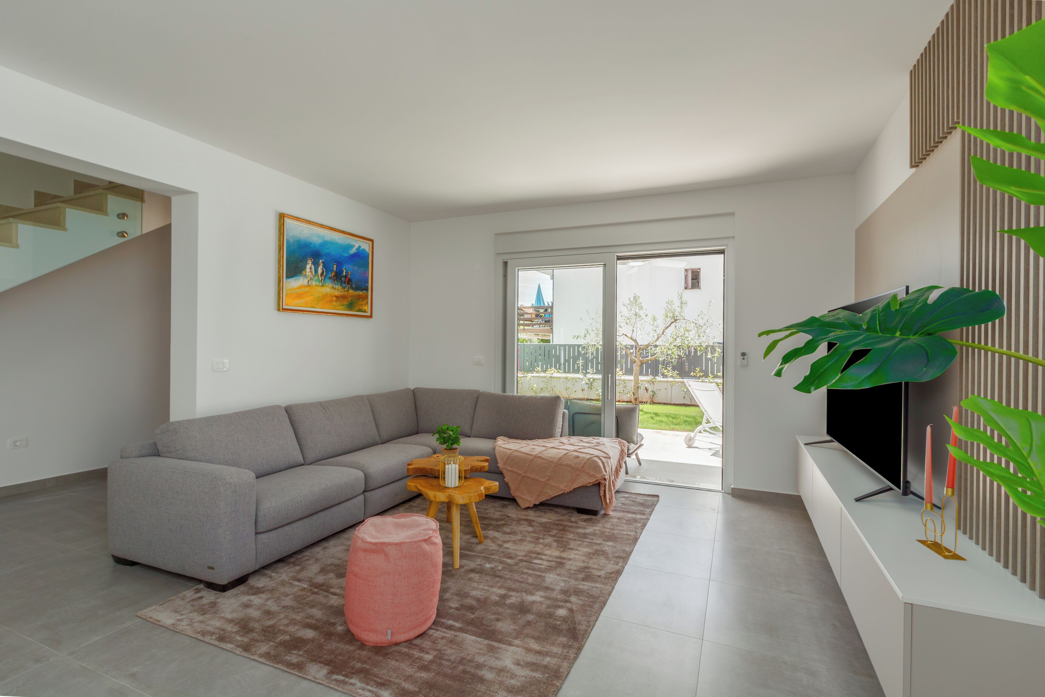 Ein modernes Wohnzimmer mit einem grauen Sofa, einem kleinen Holztisch, einem pinken Hocker und einer großen Zimmerpflanze.