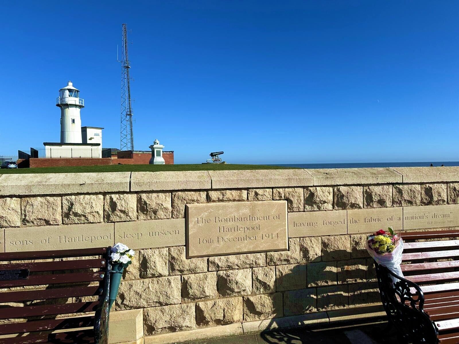 Un mur de pierre sur lequel est fixée une plaque commémorative à la mémoire de Hartlepool, un phare et un banc agrémenté d’un bouquet de fleurs.