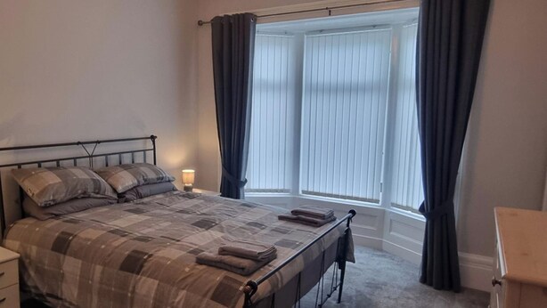Een slaapkamer met een bed, nachtkastje, dressoir en een raam met gordijnen.