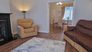 Een woonkamer met een beige fauteuil, een bruine bank en een tapijt met een marmerpatroon. Links bevindt zich een open haard, en er is een eethoek zichtbaar door een open doorgang.