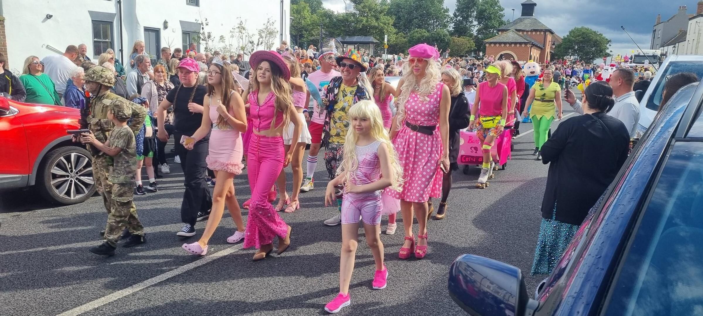 A crowded street with people walking, some in pink outfits, and a red car parked on the side.