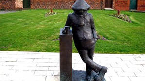 Een standbeeld van een figuur in hoed en jas, op een sokkel staand, met een gazon en een bakstenen muur op de achtergrond.
