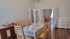 Een eetgedeelte met een lange tafel voor vier personen, omringd door houten stoelen met beklede zitplaatsen. Een buffet met manden is links zichtbaar.