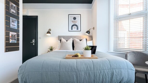 Une chambre à coucher comprenant un lit, des lampes de chevet, une œuvre d’art accrochée au mur, une porte et une fenêtre avec des stores.