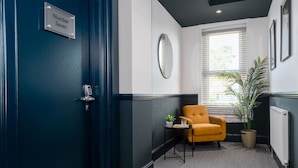 Un couloir avec une porte bleu foncé portant l’inscription « Numéro sept », une petite table avec une plante, un fauteuil jaune et une grande plante en pot près d’une fenêtre.