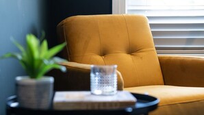 Un fauteuil de couleur moutarde, avec une petite table placée à côté, sur laquelle se trouvent un pot de plante et une carafe en verre.