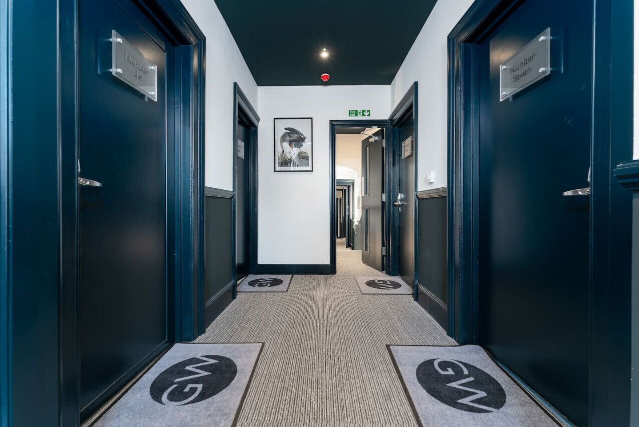 Un corridoio d'albergo con porte blu scuro, un quadro incorniciato al muro e un tappeto con un logo.