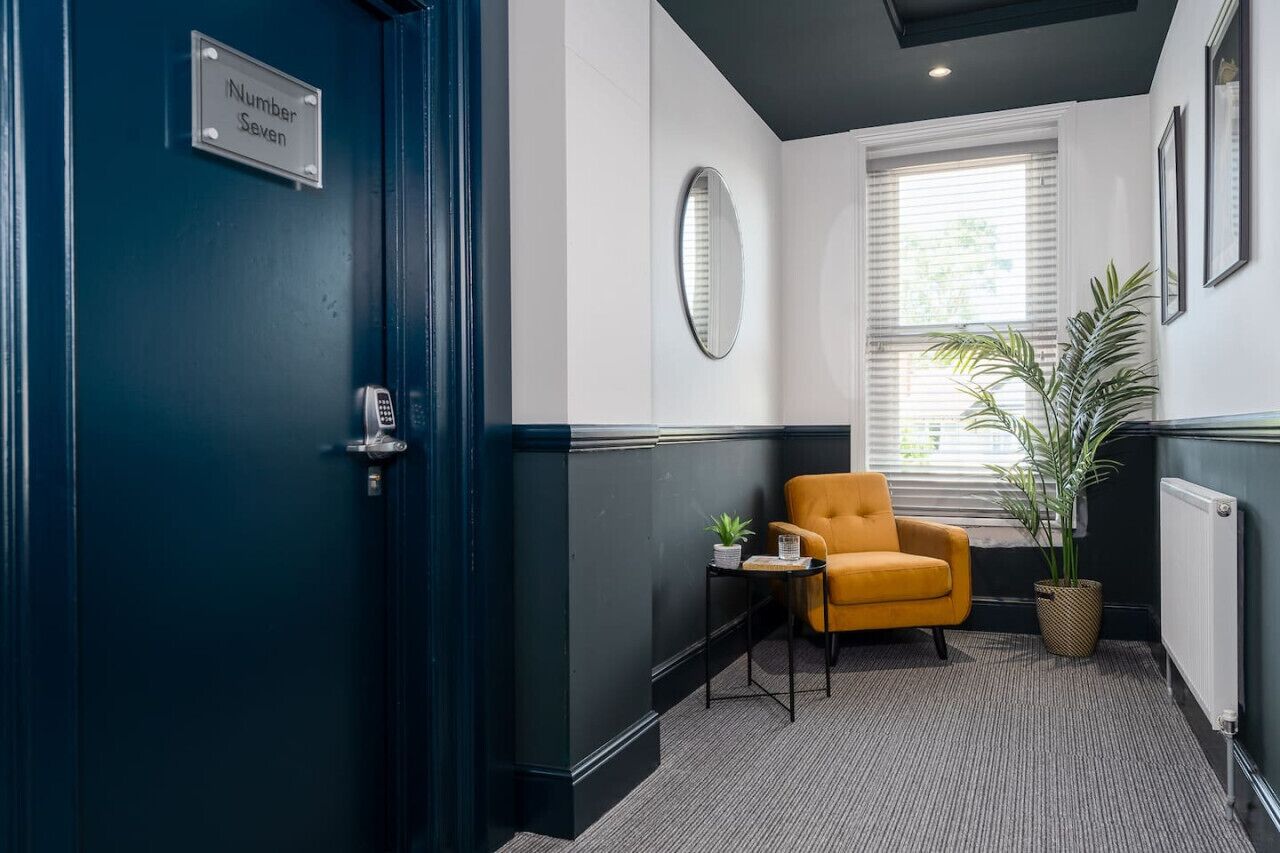 Un couloir avec une porte bleu foncé portant l’inscription « Numéro sept », une petite table avec une plante, un fauteuil jaune et une grande plante en pot près d’une fenêtre.