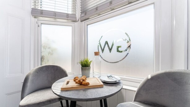 Un coin accueillant avec deux fauteuils gris, une petite table ronde, une planche à découper en bois sur laquelle sont disposés des croissants, une assiette blanche, une fourchette en argent, une petite plante en pot et une fenêtre à vitrage dépoli ornée des lettres « WCA ».