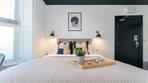 A neatly arranged hotel room with a large bed, a bedside table with a tray of breakfast items, a potted plant, and a framed artwork above the bed. - Stylish Studio; Free Laundry, Long Stay Discounts (Southampton)