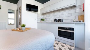 A modern bedroom with a large bed, a bedside table with a tray, a TV mounted on the wall, a built-in oven, and a kitchen area with white cabinets and a tiled backsplash. - Stylish Studio; Free Laundry, Long Stay Discounts (Southampton)