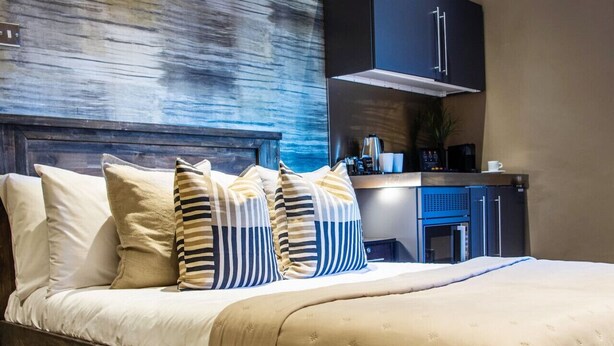 Un lit bien fait, recouvert d’une couvre-lit beige et agrémenté d’oreillers à rayures, avec une tête de lit en bois, et un coin cuisine moderne comprenant une machine à café et des placards en arrière-plan.