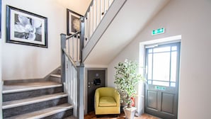 Un escalier avec une rampe grise, un fauteuil jaune, une plante en pot et une porte avec un panneau vert indiquant la sortie.