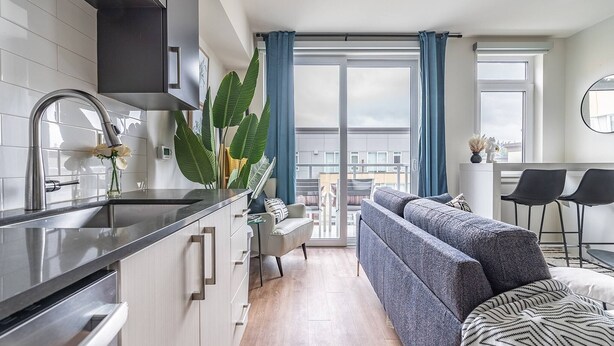 Une cuisine moderne avec un évier en acier inoxydable, un coin repas comprenant un canapé gris et une petite table, et un balcon agrémenté de rideaux bleus.