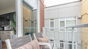 Un balcon doté d’une balustrade en métal blanc, d’un banc gris avec des coussins à rayures et d’une vue sur un immeuble moderne aux grandes fenêtres.