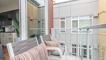 Ein Balkon mit weißem Metallgeländer, einer grauen Bank mit gestreifter Polsterung und Blick auf ein modernes Gebäude mit großen Fenstern.