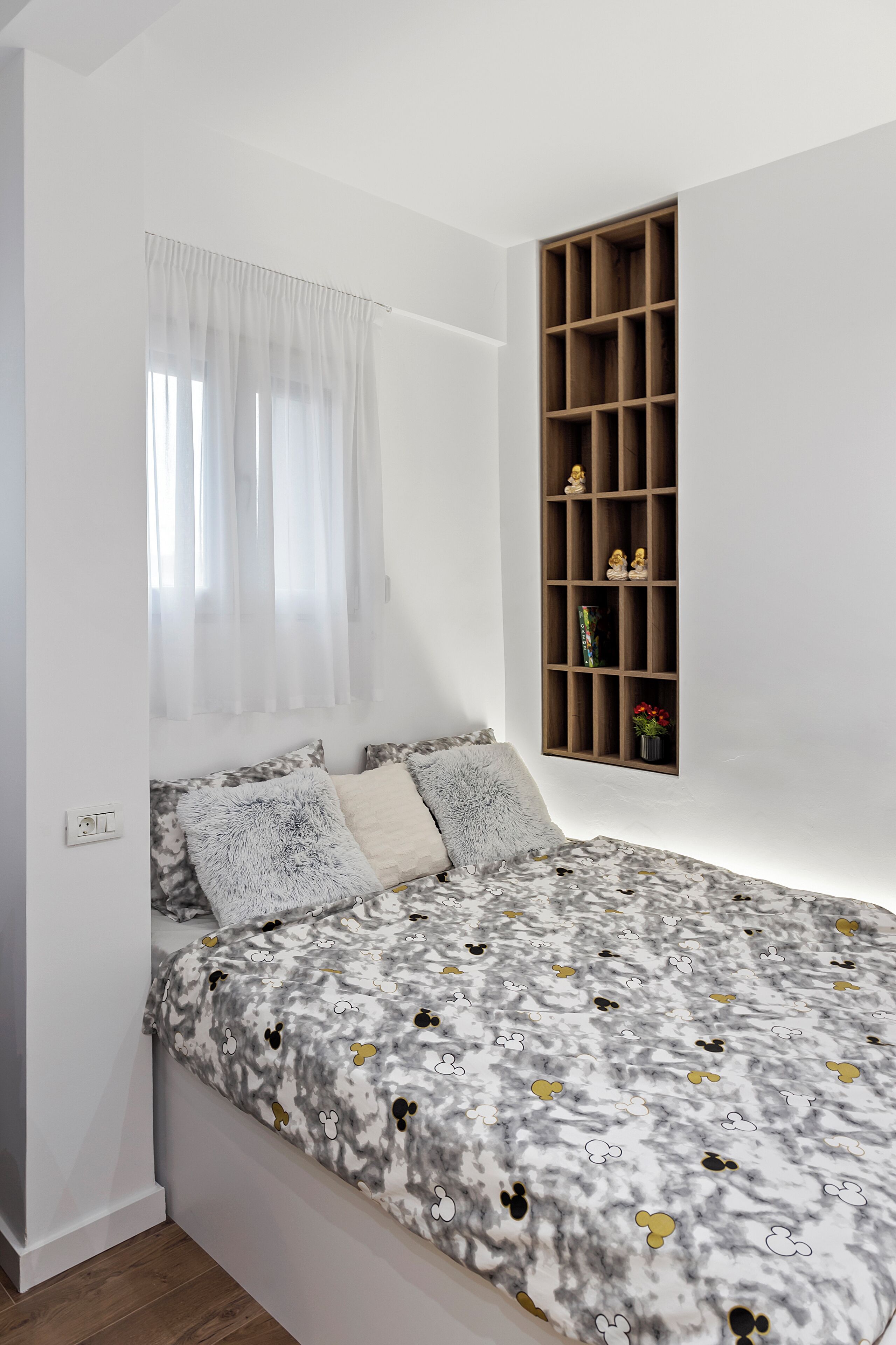 Une chambre à coucher avec un lit, une fenêtre ornée de rideaux légers, une étagère intégrée en bois agrémentée d’objets décoratifs et une couvre-lit à motifs.